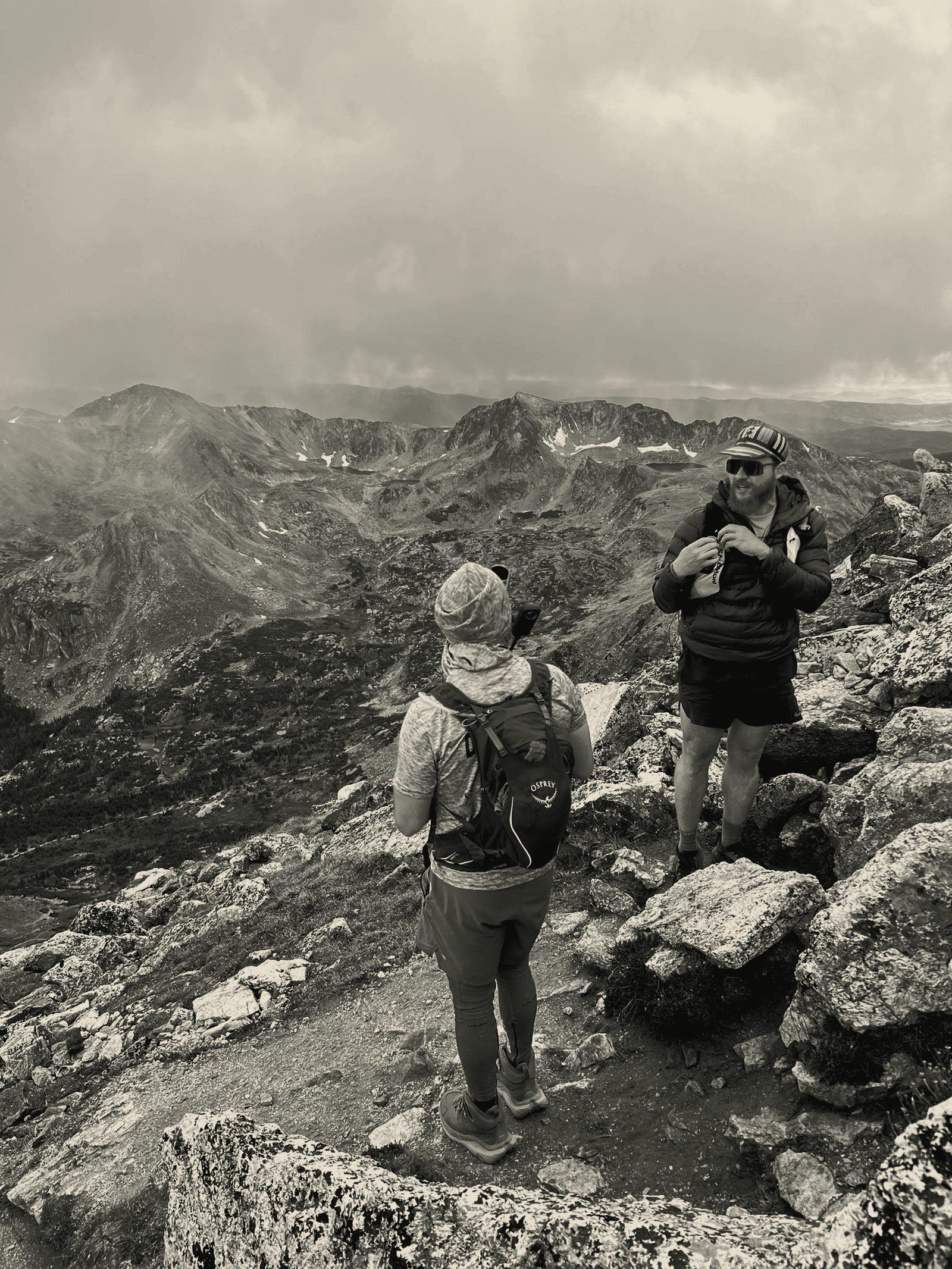 Two men on an exposed alpine ridge above a storm-lit basin, packs on, mid-expedition